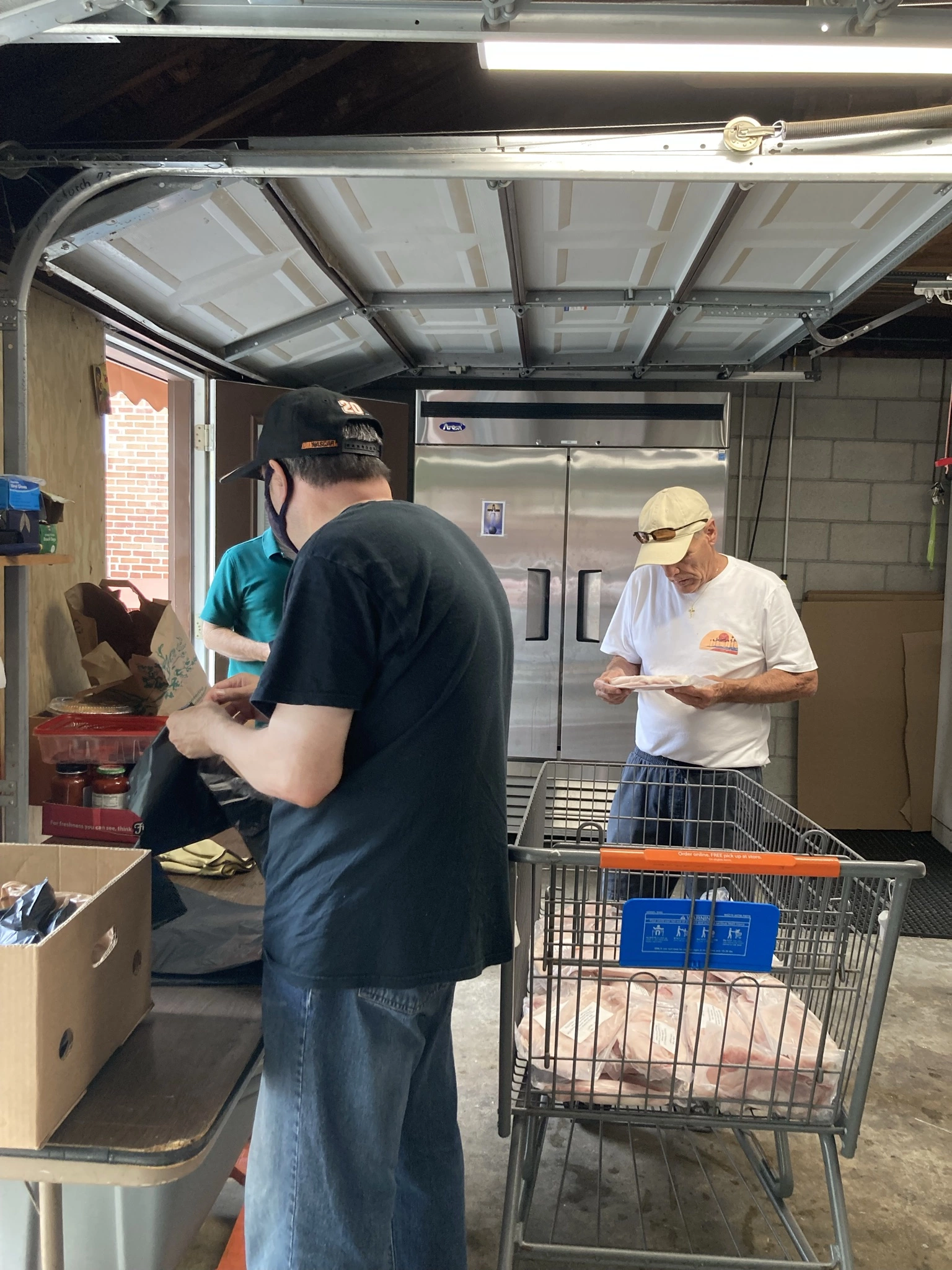 3 men in garage standing in front of refrigerator filling a shopping cart with various food items