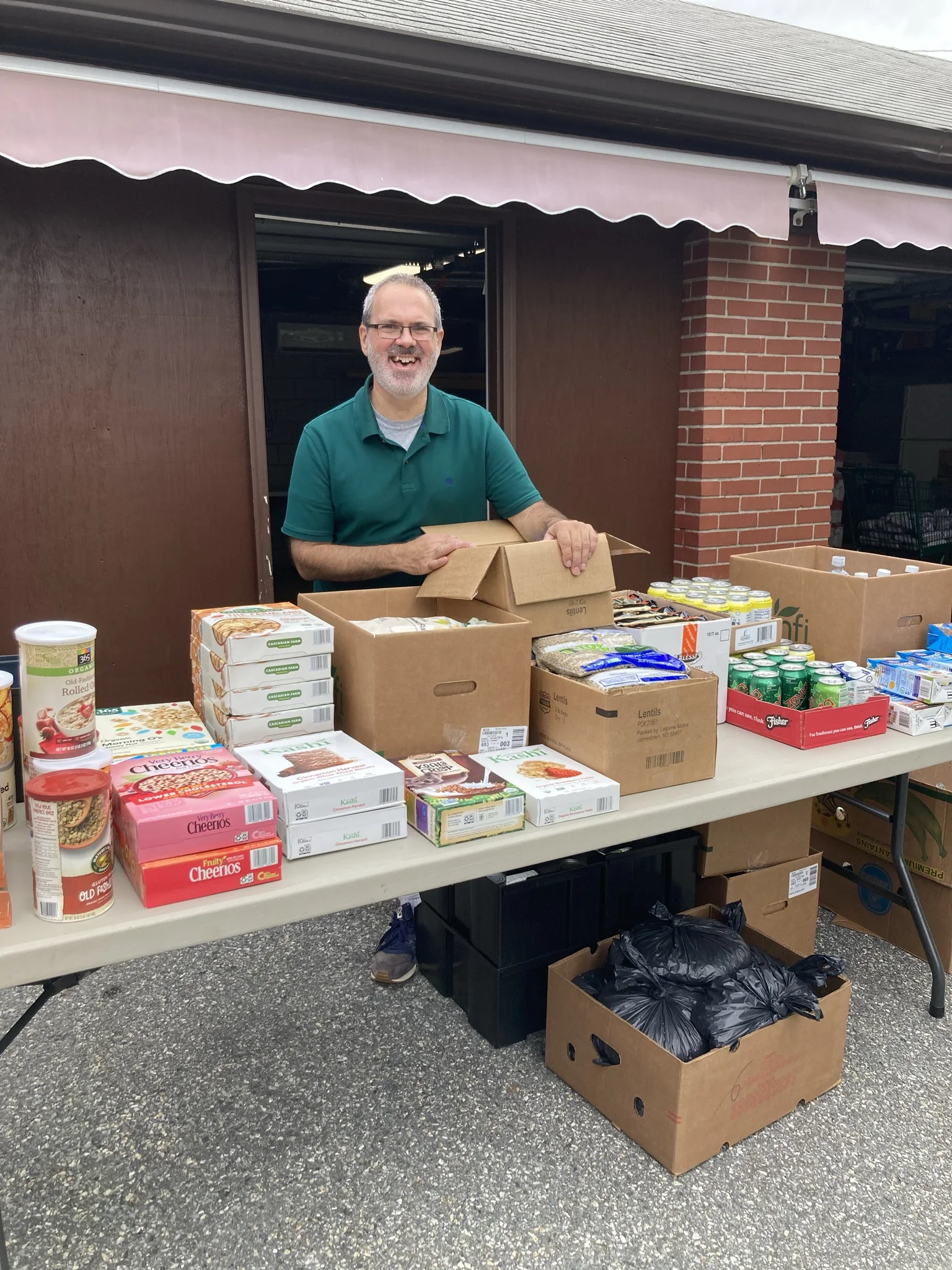 Man in green shirt standing over a table full of food in boxes like cereal, soup, and other non perishable items